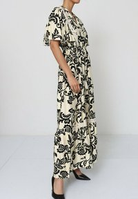 Robe maxi en crème avec un motif florale noir, dotée de manches courtes volantées et d'une taille élastiquée, se terminant par un ourlet volanté.