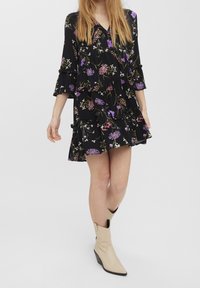 Musta kukallinen mekko, jossa on violetteja ja valkoisia kuvioita, röyhelöreunat ja hihat. Yhdistetty vaaleanbeigeihin kärkitossuihin.