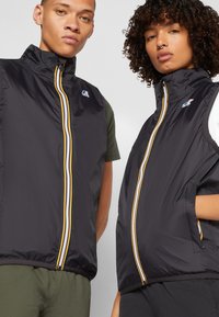 Sorte ermeløse vester med glidelås foran, hvite og gule striper langs glidelåsene, med logoer på øvre venstre side. Lett materiale.