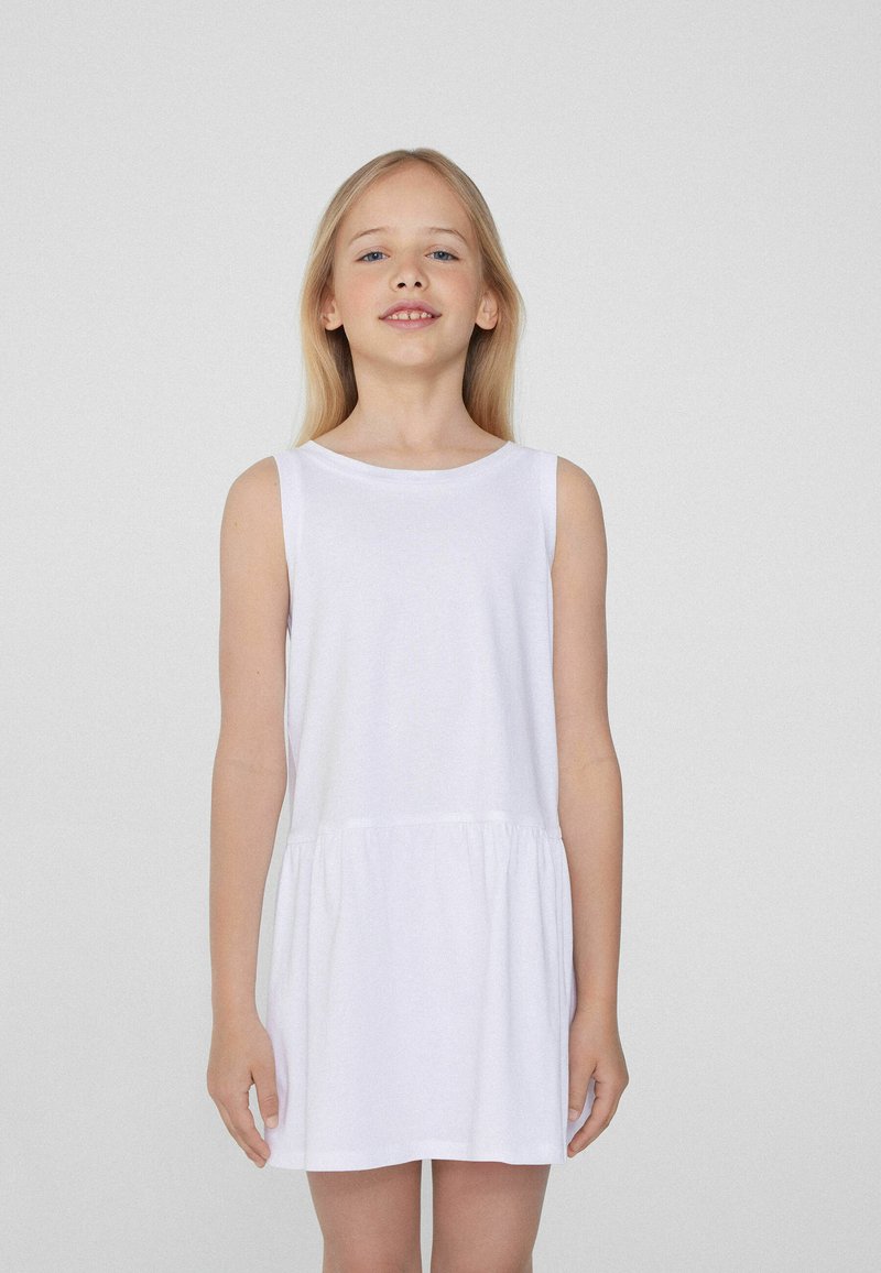 Robe blanche sans manches en tissu doux, avec un col rond et une jupe froncée, tombant au-dessus du genou, avec une texture lisse.