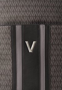 Ponovljeni geometrijski uzorak s međusobno povezanim oblikom slova "V" na teksturiranom materijalu, centrirane vertikalne crne i sive kožne pruge, srebrni "V" amblem.