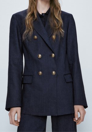 Woman wearing a dark blue double-breasted blazer with six gold buttons and matching trousers, standing against a plain background.