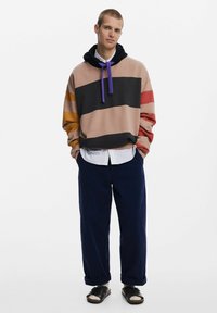 Sudadera de algodón con rayas horizontales en beige, negro, naranja y rojo. Presenta una capucha con cordón y un aspecto de capas con una camiseta blanca y pantalones azul oscuro.