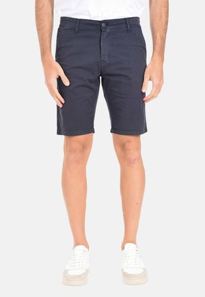 Uomo con pantaloncini casual blu navy e scarpe da ginnastica bianche, in piedi contro uno sfondo chiaro e semplice.