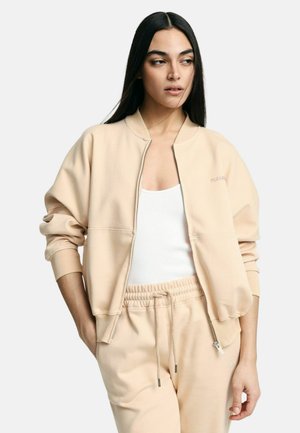 Donna con capelli lunghi e scuri che indossa una giacca beige con cerniera sopra una maglietta bianca e pantaloni della tuta beige abbinati, in piedi con una mano in tasca.