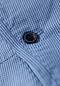 Ljusblått tyg med ett diagonalt randmönster, med en svart fyra-håls knappdetalj och synliga sömnadsdetaljer.