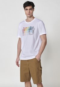 T-shirt bianco in cotone con una grafica colorata di biciclette, abbinato a pantaloni cargo beige, caratterizzati da tasche e una vestibilità rilassata.