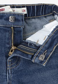 Jeans dżinsowe w ciemnoniebieskim kolorze z złotym suwakiem i zapięciem na guzik. Posiadają wyszywaną talię oraz wewnętrzną podszewkę widoczną w pobliżu suwaka.