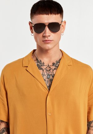 Chemise à manches courtes en tissu jaune moutarde, dotée d'un col, d'un devant à boutons et d'une texture lisse. Lunettes de soleil noires et colliers en argent superposés.