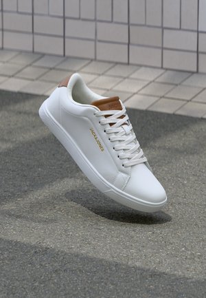 Baskets en cuir blanc avec des accents bruns, lacets noués, marquées "JACK & JONES" sur le côté et sur la languette, montrées flottant au-dessus de l'asphalte près d'un mur carrelé.