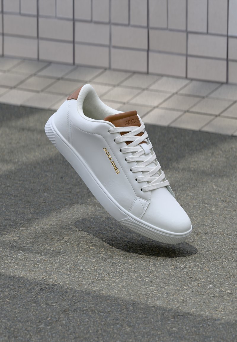 Baskets en cuir blanc avec des accents bruns, lacets noués, marquées "JACK & JONES" sur le côté et sur la languette, montrées flottant au-dessus de l'asphalte près d'un mur carrelé.