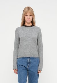 Grijze gebreide trui met een ronde halslijn en kantafwerking. Lange mouwen en een relaxte pasvorm. Gepaard met high-waisted blauwe jeans.