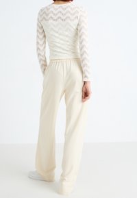 Krémově zbarvený krajkový top se vzorem cikcak, spojený s kalhotami v béžové barvě s vysokým pasem a širokými nohavicemi. Oba kusy mají měkkou texturu a volný střih.