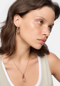 Ohrringe aus Roségold mit ineinander verwobenen Designs und einer passenden Anhängerhalskette an einem Modell mit mittelbraunem Haar, das ein helles Tanktop trägt.