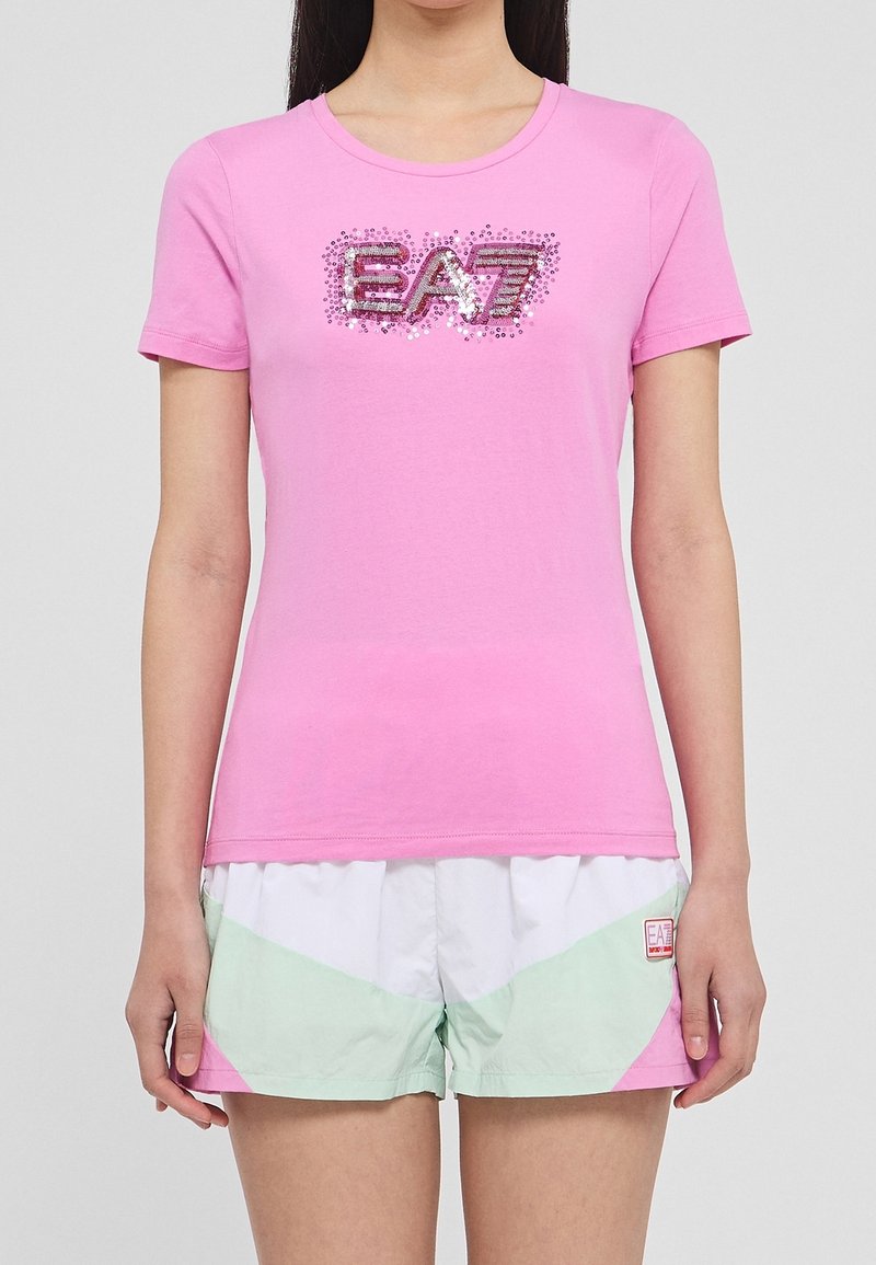 Růžové bavlněné tričko s kulatým výstřihem a třpytivým logem "EA7". Kombinováno s pastelemi zelenými a bílými šortkami se geometrickými vzory.