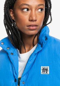 Blue lightweight jacket with a textured finish, snap buttons, and a logo patch on the upper left. Worn over a light gray shirt.