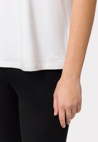T-shirt bianco con orlo arrotondato e un piccolo logo "LES LUMES" in basso a sinistra, abbinato a leggings neri, che mettono in evidenza una texture di tessuto liscia.