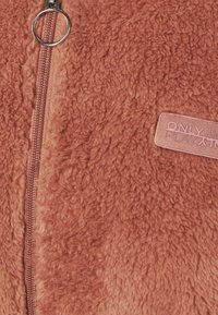 Chaqueta de forro polar en naranja quemado, con una superficie texturizada, cremallera plateada y una etiqueta cosida que dice "SOLO JUEGO". Diseño suave y cálido.