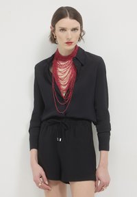 Donna che indossa una camicia nera con bottoni e pantaloni corti, con una collana di perline rosse stratificate e rossetto rosso, su sfondo bianco.