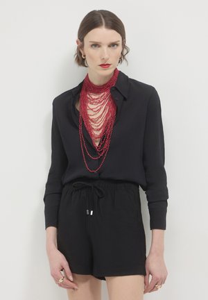 Woman wearing a black buttoned shirt and shorts with layered red beaded necklace and red lipstick against white background.