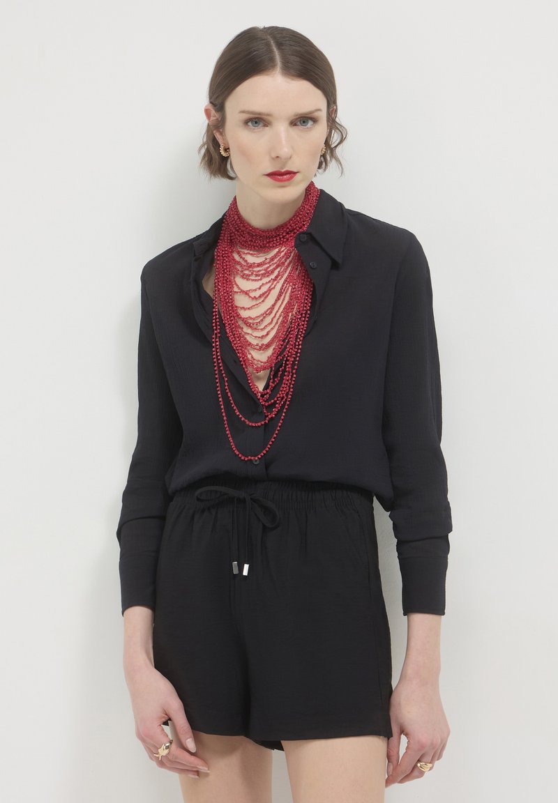 Donna che indossa una camicia nera con bottoni e pantaloni corti, con una collana di perline rosse stratificate e rossetto rosso, su sfondo bianco.