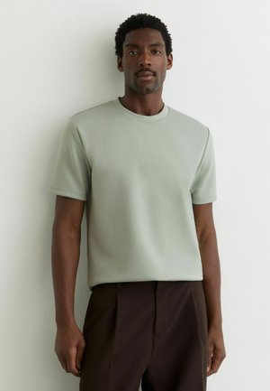 Bărbat cu păr scurt și creț, purtând o cămașă verde pal cu mâneci scurte și pantaloni maro închis, stând în picioare lângă un perete alb.