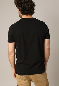 Man met krullend haar in een zwart T-shirt met korte mouwen en beige cargobroek, staande met de rug naar de camera tegen een neutrale achtergrond.