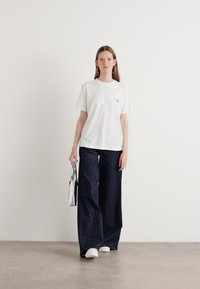 White cotton t-shirt with small logo, paired with dark denim wide-leg pants. Model holds a silver bag and wears white shoes.