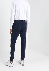 Person som bär marinblå joggingbyxor med "Just Do It"-text på vänster ben, vit långärmad skjorta och vita Nike-sneakers, står vänd bort.