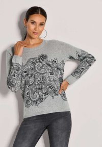 Frau mit dunklem Haar im Pferdeschwanz, die große Creolen trägt und einen grauen Pullover mit komplizierten schwarzen floralen Paisley-Mustern trägt, posiert mit einer Hand auf der Hüfte.
