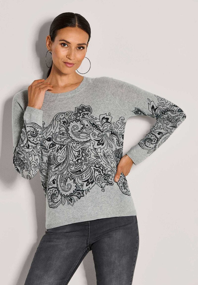 Frau mit dunklem Haar im Pferdeschwanz, die große Creolen trägt und einen grauen Pullover mit komplizierten schwarzen floralen Paisley-Mustern trägt, posiert mit einer Hand auf der Hüfte.