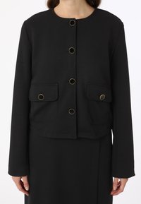 Veste noire raccourcie en tissu lisse, avec un col rond, des boutons en ton doré et deux poches à rabat à l'avant.