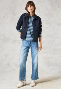 Marineblaues Zip-Hoodie über einem blau-grün gestreiften Hemd, kombiniert mit hellblauen weit geschnittenen Jeans und weißen Sneakern mit schwarzen Akzenten.