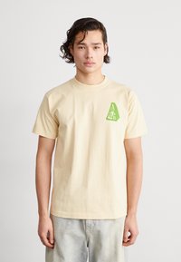 Camiseta de algodón beige de manga corta con un logotipo geométrico verde en la parte izquierda del pecho. El modelo lleva pantalones de mezclilla claros.