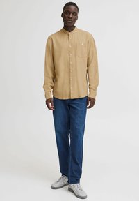 Lys beige langermet skjorte med rund krage, som har en brystlomme og knappelukking, kombinert med mørkeblå jeans og grå joggesko.