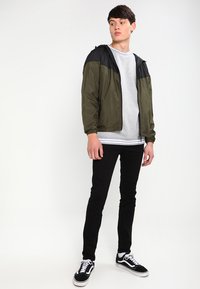 Let jakke med olivengrønne og sorte paneler, lynlås foran, parret med en grå skjorte og sorte skinny jeans. Sorte og hvide sneakers.