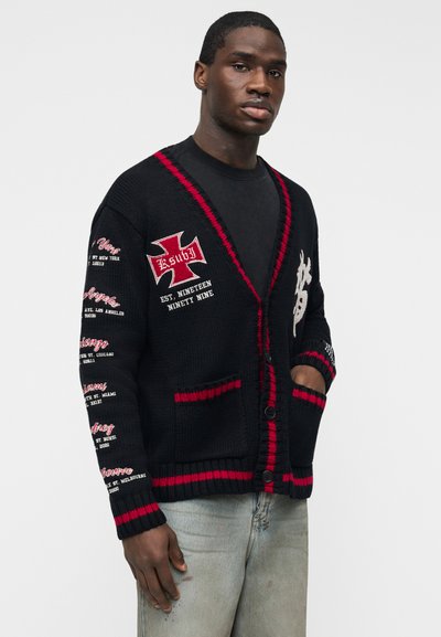 Uomo che indossa un cardigan nero lavorato a maglia con bordi rossi e toppe di testo bianche, abbinato a jeans azzurri, in piedi davanti a uno sfondo semplice.