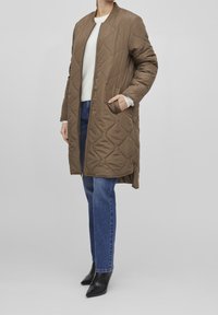 Brun vadderad lång kappa med rund halslinning, dragkedja fram, och sidofickor, kombinerad med blå jeans och svarta spetsiga skor.