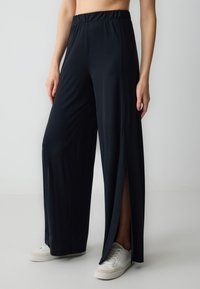 Pantaloni neri a gamba larga con vita elastica, caratterizzati da aperture laterali e tessuto leggero e morbido. Abbinati a sneaker bianche.