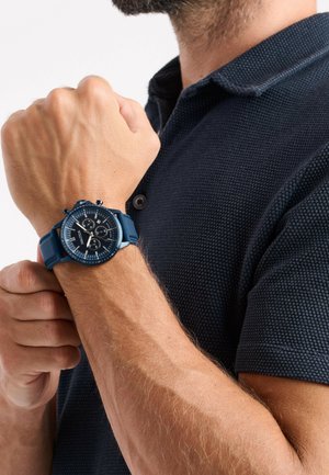 Orologio cronografo blu con quadrante nero rotondo, cinturino in silicone blu con trama, indici delle ore prominenti e tre sottodial, indossato su un polso.