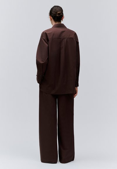 Personne avec les cheveux attachés en chignon, portant une chemise ample à manches longues marron foncé et un pantalon large assorti, debout devant un arrière-plan uni.
