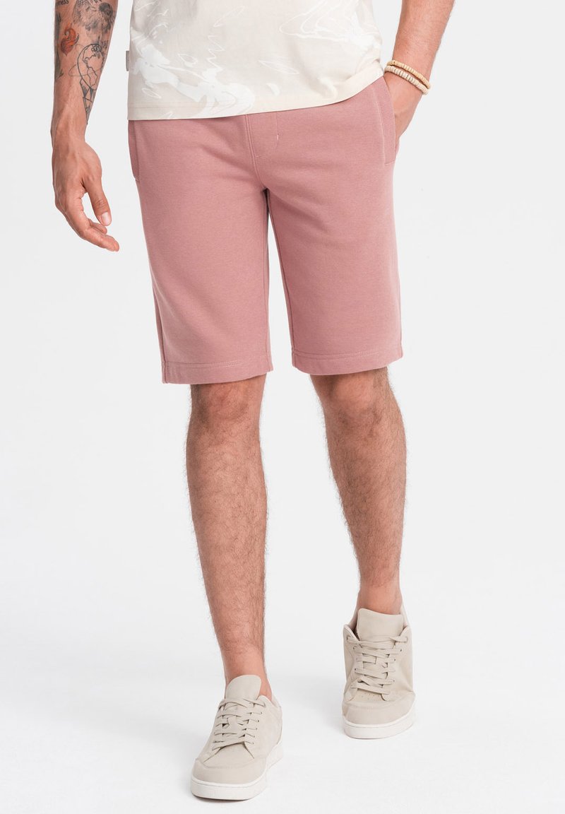 Světle růžové bavlněné šortky s elastickým pasem, mají boční kapsy a rovný střih. Na sobě má béžové tenisky.