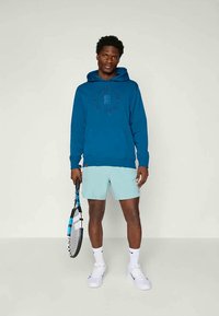 Homem a usar um hoodie azul, calções azuis claros, ténis brancos e meias, segurando uma raquete de ténis, em pé contra um fundo simples.