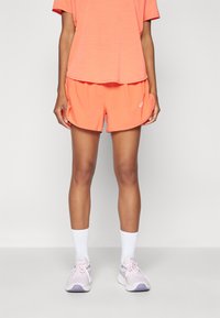 Conjunto deportivo de mujer que incluye una camiseta corta y suelta de color naranja brillante y pantalones cortos a juego, con calcetas blancas hasta el tobillo y zapatillas de deporte de color rosa claro.