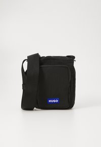 Svart crossover-veske laget av teksturert nylon, med en frontlomme og et blått "HUGO" logo-patch. Justerbar stoffrem for enkelhet.