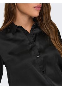 Blouse en satin noir dotée d'un col classique, d'une patte de boutonnage à l'avant et d'une texture lisse, avec un léger éclat et sans motifs ni accents visibles.