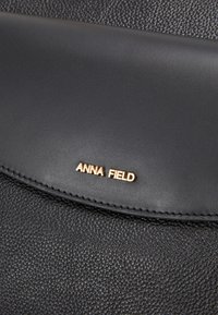 Sac à main en cuir noir présentant une surface texturée, un rabat lisse et un logo embossé doré "ANNA FIELD". Les accents incluent des coutures détaillées.