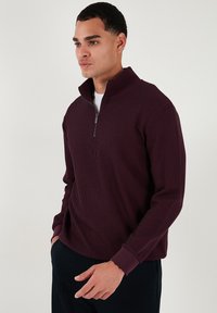 Suéter de color burdeos con canalé, con cuello alto con cremallera, mangas largas y puños en contraste. Textura suave, diseñado para un ajuste ceñido.