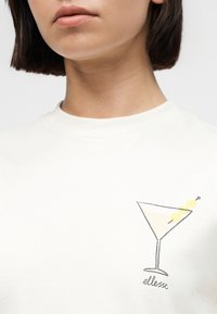 Κοντινό πλάνο ενός ατόμου που φοράει λευκό πουκάμισο με μικρό σχέδιο μαρτίνι και ελιές στο στήθος.