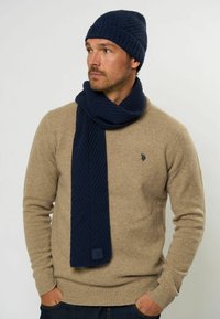 Marineblå strikket hue og tørklæde sæt med en tekstureret overflade, parret med en beige sweater. Tørklædet har et logo patch.